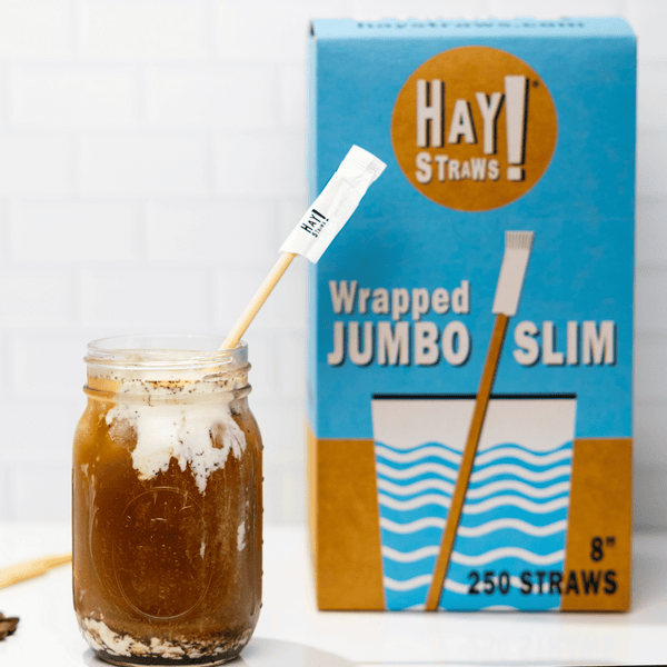 HAY Straws Wrapped Standard Drinking Straws made from reed plant stems, Individually wrapped straws, a biodegradable straw in an iced coffee with a wrapped Jumbo slim straw box in the background, 8” length, non soggy straws, compostable straws, natural straws