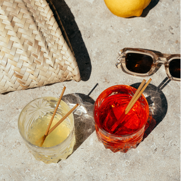 HAY! Straws Cocktail Straws in two lowball cocktail crystal glasses on a natural travertine stone table, with sunglasses and a straw bag. It's an elegant scene cocktail straws