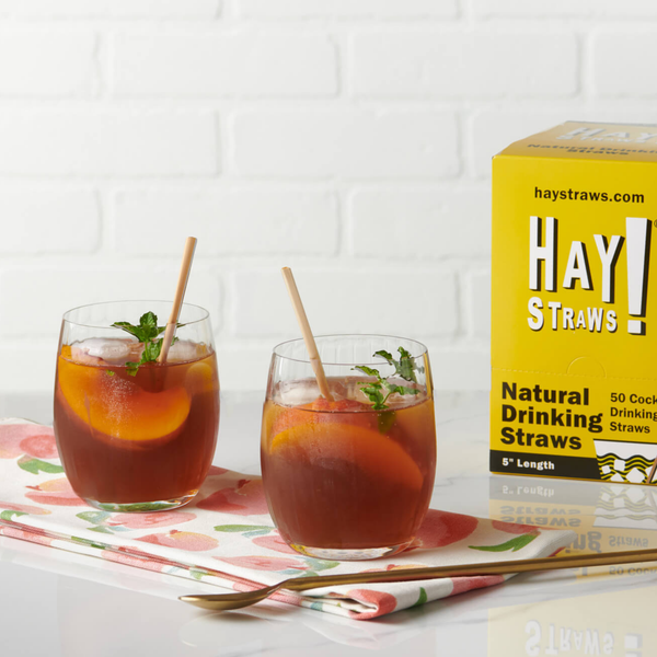 Two glasses of iced tea with hay straws 5" straws inside the lowball glass on a table next to a box of hay straws with fresh peaches and mint, natural and compostable drinking straws