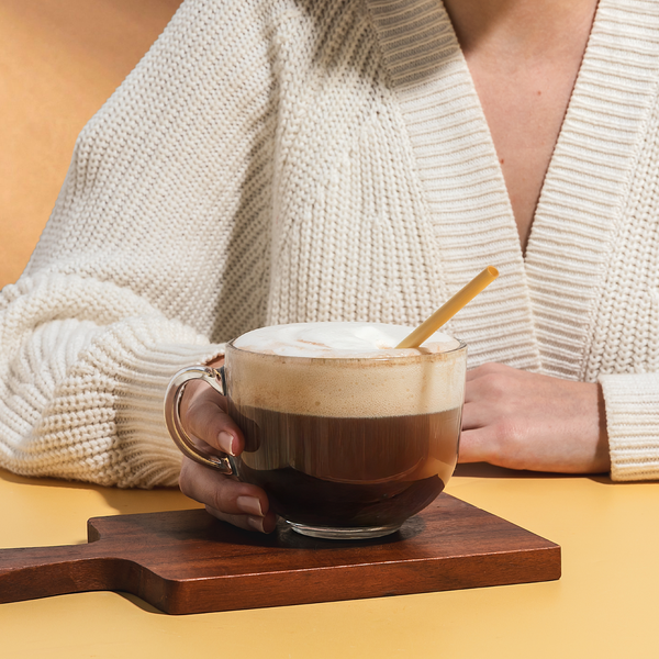 Woman holding a coffee cup with a jumbo slim 6" straw by HAY! Straws on a wooden coaster on a yellow surface