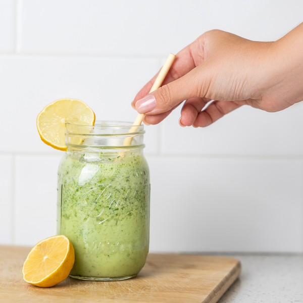 Green smoothie in a jar with a lemon slice on a wooden board and a woman's hand holding a plastic-free HAY! straw made from reed stems and looking like a bamboo straw