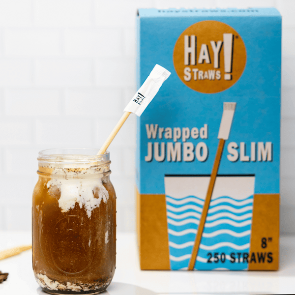 HAY Straws Jumbo Slim Straws made from reed plant stems, Individually wrapped straws, a biodegradable straw in an iced coffee with a wrapped Jumbo slim straw box in the background, 8” length, non soggy straws, compostable straws, natural straws