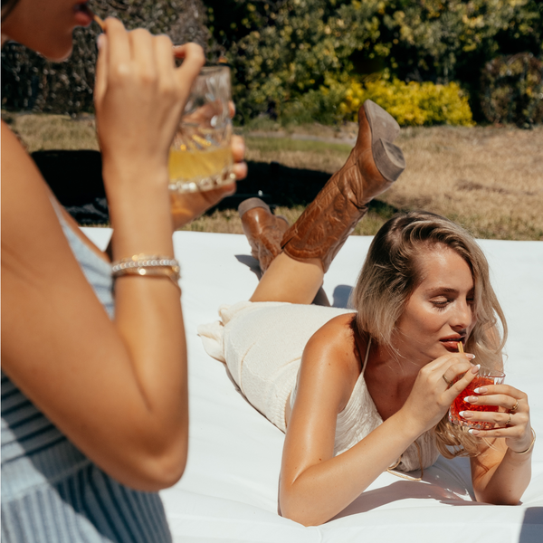 Two women lounging outdoors, one drinking from a glass and the other from a red cup. Sipping their cocktial using HAY! straws original 5" cocktail straw
